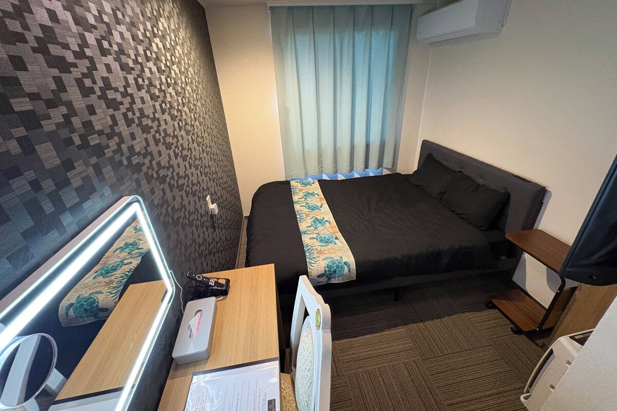 A hotel room with a bed, a desk with a mirror, a remote control, a tissue box, and a window with curtains.