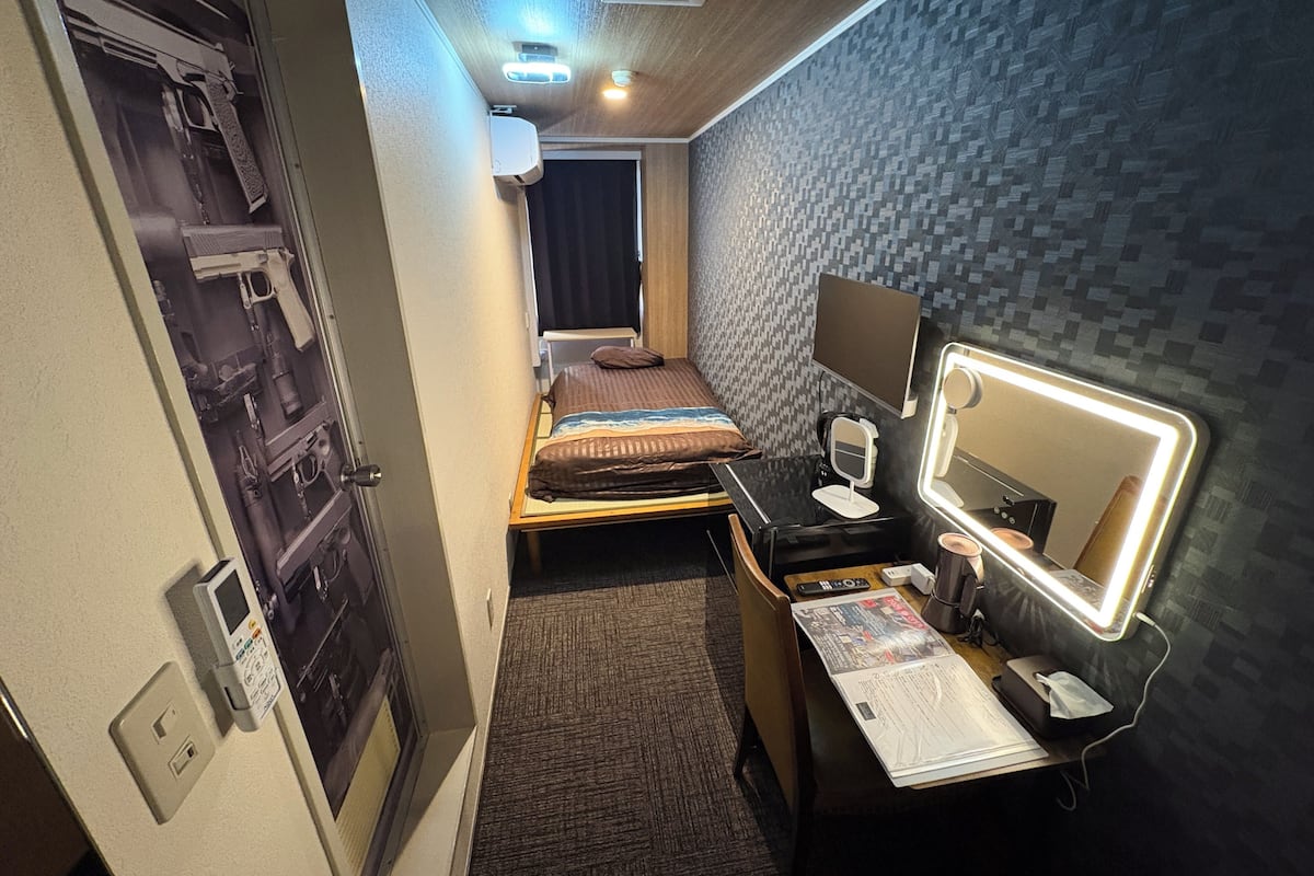 A compact hotel room with a bed, bedside table, mirror, and a small desk with a lamp.