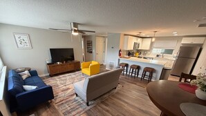 Un salon moderne avec un canapé, un fauteuil et une table à manger avec des chaises. On aperçoit, au fond, une cuisine avec un coin bar.