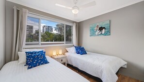 A bedroom with two beds, a window with curtains, a ceiling fan, and a painting of a pelican. - Seychelle unit 5 Kings Beach (Kings Beach)