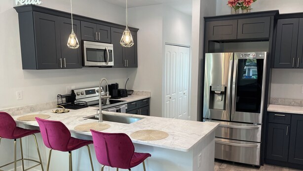 A modern kitchen with dark cabinets, a stainless steel refrigerator, a microwave, and a large island with bar stools. - The Oasis on Waters -New Home (Tampa)