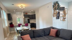 A modern living room with a sectional sofa, a dining area with two red chairs, and a kitchen in the background. - The Oasis on Waters -New Home (Tampa)