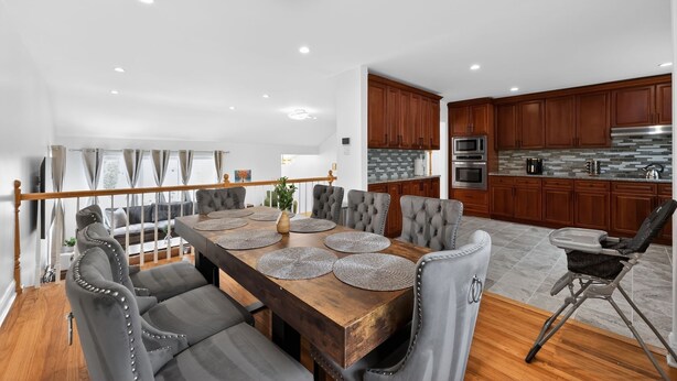 A modern kitchen with wooden cabinets, a dining area with a large wooden table, and grey upholstered chairs.