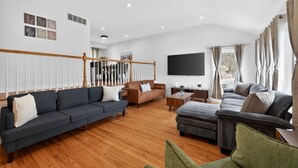 A spacious living room with a wooden floor, a grey sofa, a brown sofa, a grey sectional, and a TV mounted on the wall.