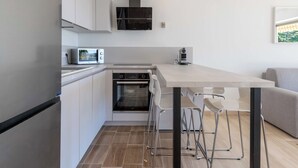 Une cuisine moderne équipée d’un four encastré, d’un micro-ondes et d’une machine à café. On y trouve une table à manger avec des chaises, ainsi qu’un canapé dans le salon attenant.