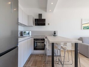 A modern kitchen with a built-in oven, microwave, and a coffee machine. There is a dining table with chairs and a sofa in the adjacent living area. - 1-Br Sea-View Apartment – Prestigious Residence – Parking (Vallauris)