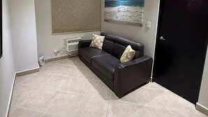 Et værelse med en sort lædersofa, beige fliser, et loft med et struktureret mønster, en vægmonteret aircondition, et vindue med en beige rullegardin og en sort dør.