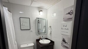 Et badeværelse med toilet, vask og spejl. Der er en bøjle til håndklæder og et indrammet citat på væggen.