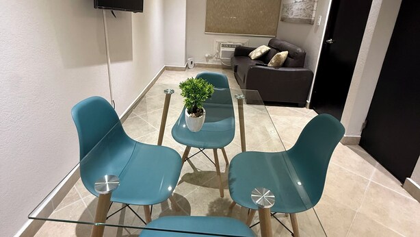 En moderne stue med et spisebord med glasplade, to blå stole, en sofa, et fjernsyn monteret på væggen og en potteplante.