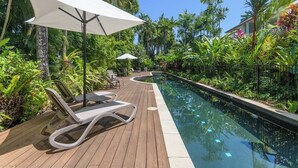 Eine Holzterrasse mit Liegestühlen und Sonnenschirmen, ein Schwimmbecken und üppige tropische Bepflanzung.