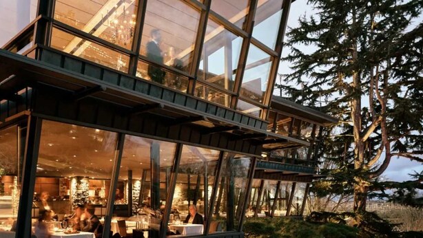 A modern building with large glass windows, a dining area with tables and chairs, and a view of trees. - Green Lake's 2B Lower Unit with Parking (Seattle)