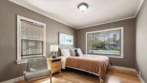 Een slaapkamer met een bed, een stoel, een nachtkastje, een lamp en twee ramen met jaloezieën.