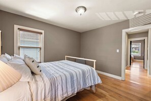 Une chambre à coucher avec un lit, une fenêtre avec des stores, un luminaire au plafond et un plancher en bois.