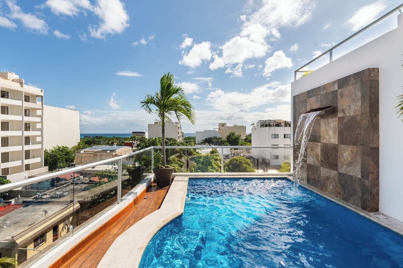 A rooftop pool with a waterfall feature, surrounded by a glass railing, and overlooking a cityscape.