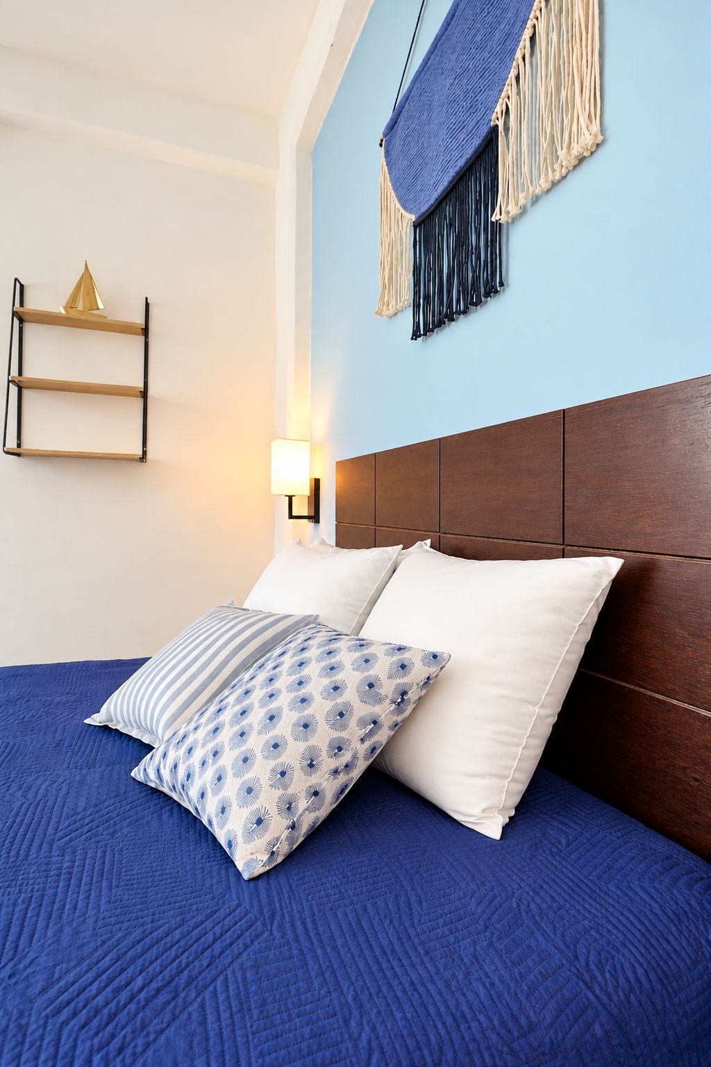 Ein Bett mit blauer Steudecke und gemusterten Kissen, ein Holz-Kopfteil, ein an der Wand befestigtes Regal und eine Wandlampe.