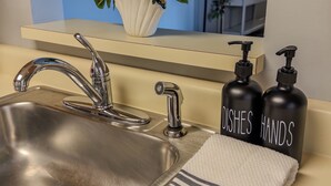Ein modernes Badezimmer mit einem Edelstahlwaschbecken, einem Blumentopf und schwarzen Seifenspendern, auf denen 'GEFLÄSCH HANDE' steht.