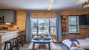 A cozy cabin interior with a wooden dining table set for four, a kitchen area with a microwave, and a bed with a quilt. - 2 Condos on Ausable River w/ Dock & Backyard (Wilmington)