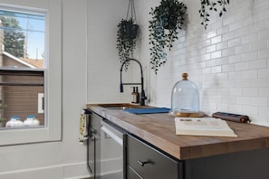 A kitchen with a window, white tiled backsplash, hanging plants, a coffee maker, and a kitchen scale. - 2 Condos on Ausable River w/ Dock & Backyard (Wilmington)
