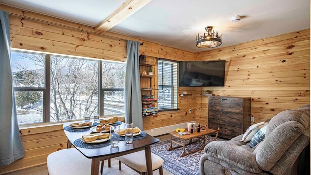 A cozy cabin interior with a dining table set for two, a sofa, a television, and a bookshelf. - 2 Condos on Ausable River w/ Dock & Backyard (Wilmington)