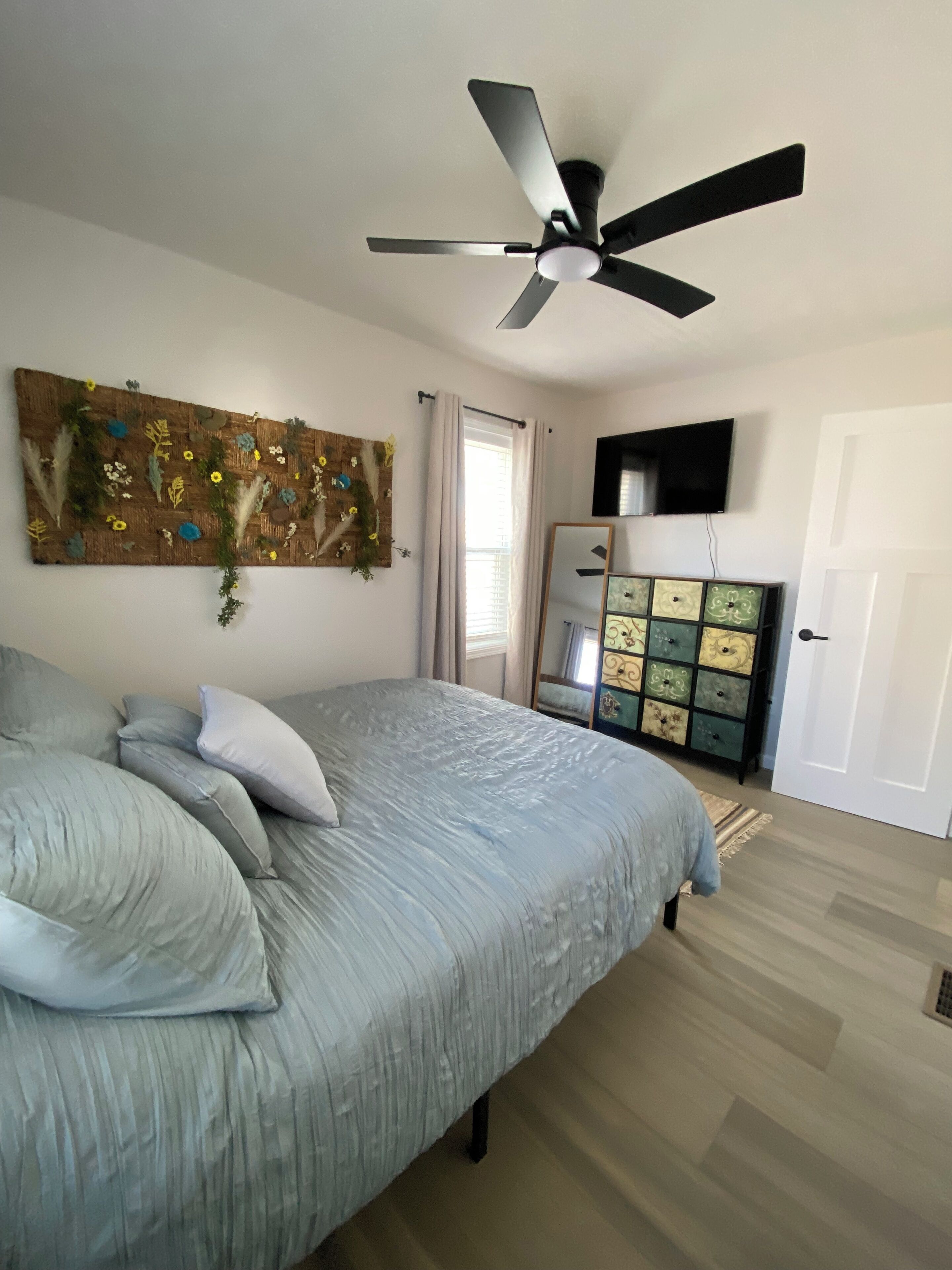 A bedroom with a bed, a ceiling fan, a TV, a dresser, and a window with curtains.