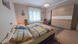 Un dormitorio con cama, una silla, una mesita, un armario y una ventana con cortinas.
