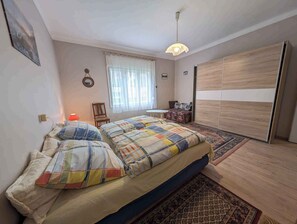 A bedroom with a bed, a chair, a small table, a wardrobe, and a window with curtains.