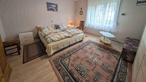 Un dormitorio con una cama, una mesita de noche, una silla, una pequeña mesa, una alfombra y una ventana con cortinas.
