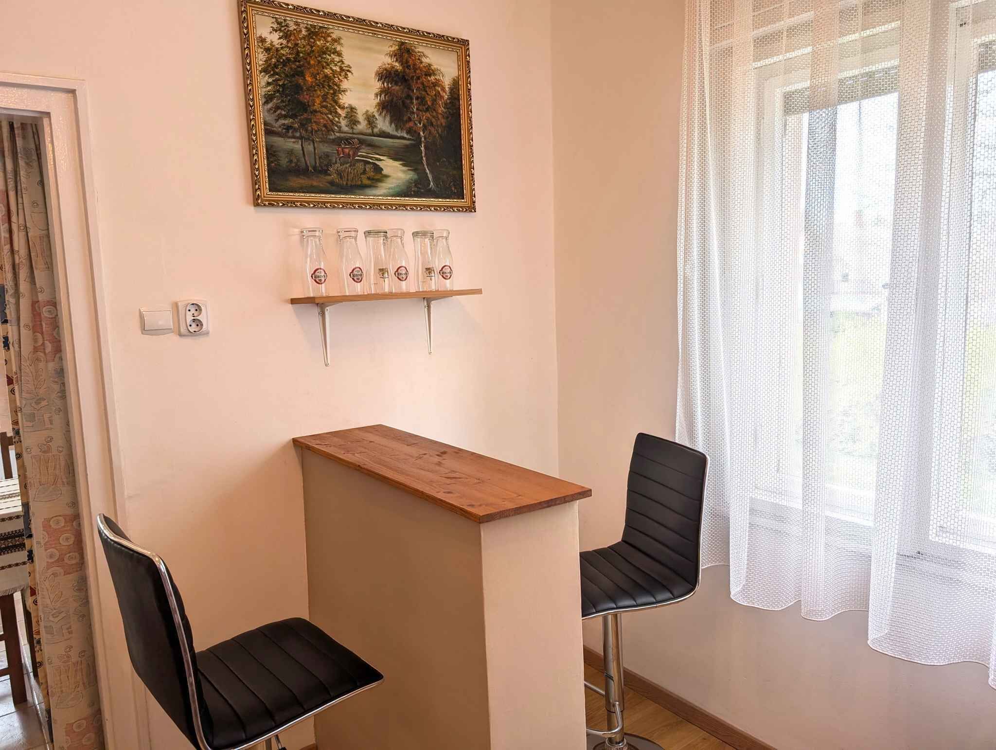 A room with a wooden table, two black chairs, a painting, and a shelf with glasses.