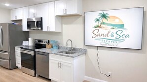 Una cocina moderna con armarios blancos, electrodomésticos de acero inoxidable, encimera de granito y un televisor empotrado mostrando 'Sand & Sea Escapes Myrtle Beach, SC'.