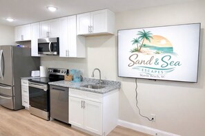 A modern kitchen with white cabinets, stainless steel appliances, a granite countertop, and a wall-mounted TV displaying 'Sand & Sea Escapes Myrtle Beach, SC'. - Full House • Walk to Beach • Families & Groups • Patio & Fire Pit • Parking (Myrtle Beach)