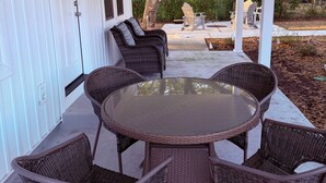 Un patio cubierto con muebles de mimbre, una mesa de vidrio y vista a una casa verde al fondo.