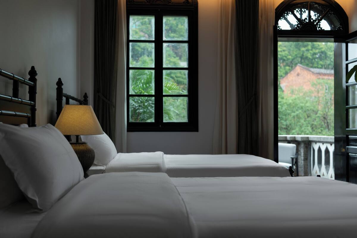 A hotel room with two beds, a window with a view of greenery, and a lamp on the nightstand.