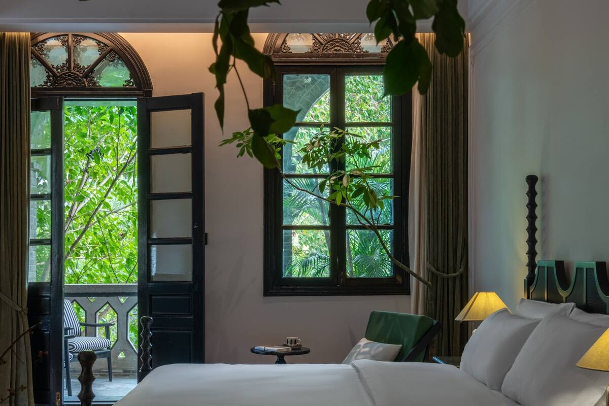 A hotel room with a large bed, two bedside tables, a chair, a window with a view of greenery, and a door leading to an outdoor area.