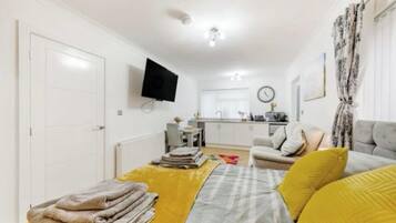 Ett kompakt bostadsutrymme med en säng, en soffa, en platt-TV, en köksdel och ett matbord.