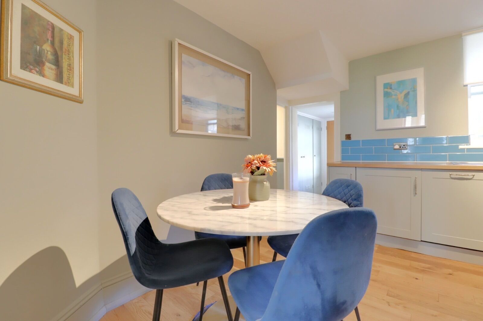Een eetgedeelte met een ronde tafel, blauwe stoelen en een bloemenvaas. Achtergrond: een keuken met blauwe tegels en witte kasten.