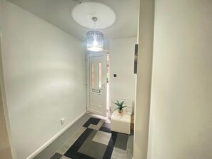 Un couloir avec un sol à carreaux, une porte blanche, un luminaire au plafond et une petite plante posée sur une table blanche.