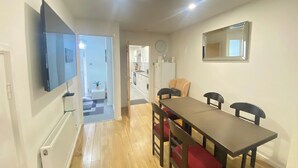 Een eetgedeelte met een tafel en stoelen, een aan de muur bevestigde televisie, een radiator en een keuken op de achtergrond.