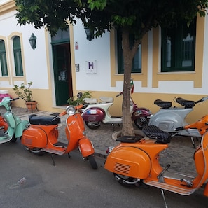 Una hilera de scooters naranja antiguos aparcados en una calle empedrada, con un árbol en el centro, y un edificio de fachada blanca y contraventanas verdes.