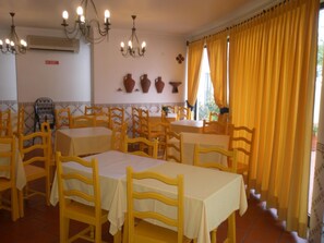 A dining area with multiple tables and chairs, an air conditioning unit, decorative wall pieces, and yellow curtains. - Hotel Passagem Do Sol (Moura)