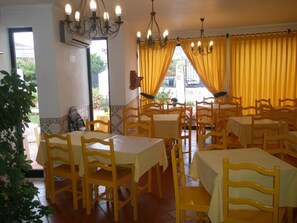 Un espace repas comprenant plusieurs tables et chaises, des rideaux jaunes et un lustre.