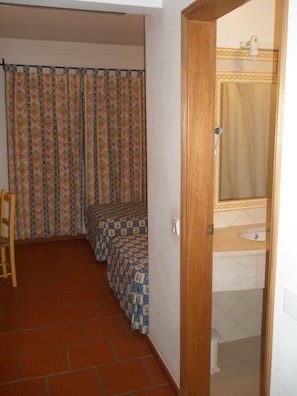 A room with a tiled floor, a bed with patterned bedding, a wooden chair, and a bathroom with a sink and mirror. - Hotel Passagem Do Sol (Moura)