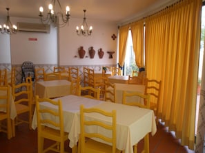 Un comedor con sillas y mesas amarillas, una araña y un ventanal con cortinas amarillas.