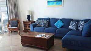 A living room with a blue sofa, a wooden coffee table, a wicker chair, and a small side table with a lamp. - Above The Dunes (Woorim)