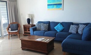 A living room with a blue sofa, a wooden coffee table, a wicker chair, and a small side table with a lamp. - Above The Dunes (Woorim)