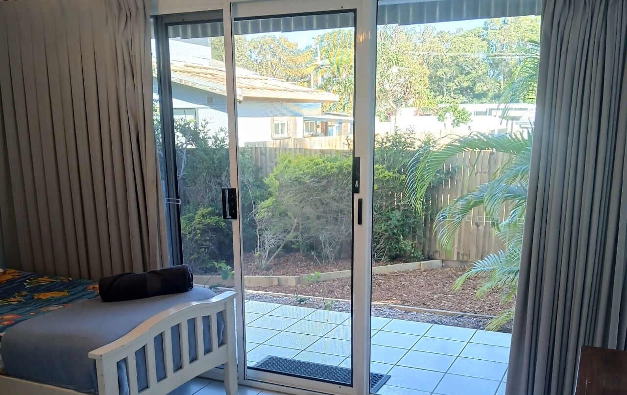 A room with a bed, a sliding glass door to a patio, and a view of a backyard with a wooden fence and greenery.