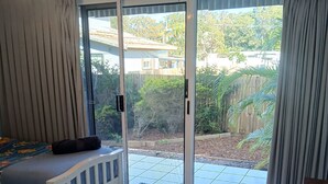 A room with a bed, a sliding glass door to a patio, and a view of a backyard with a wooden fence and greenery. - Above The Dunes (Woorim)