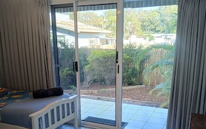A room with a bed, a sliding glass door to a patio, and a view of a backyard with a wooden fence and greenery. - Above The Dunes (Woorim)
