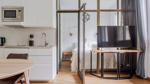 Un espacio habitable compacto con kitchenette, comedor, televisor sobre un soporte y puerta corrediza de vidrio que da a un dormitorio.