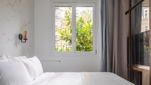 Un dormitorio con cama, ventana con vista a vegetación, lámpara de pared y armario oscuro.