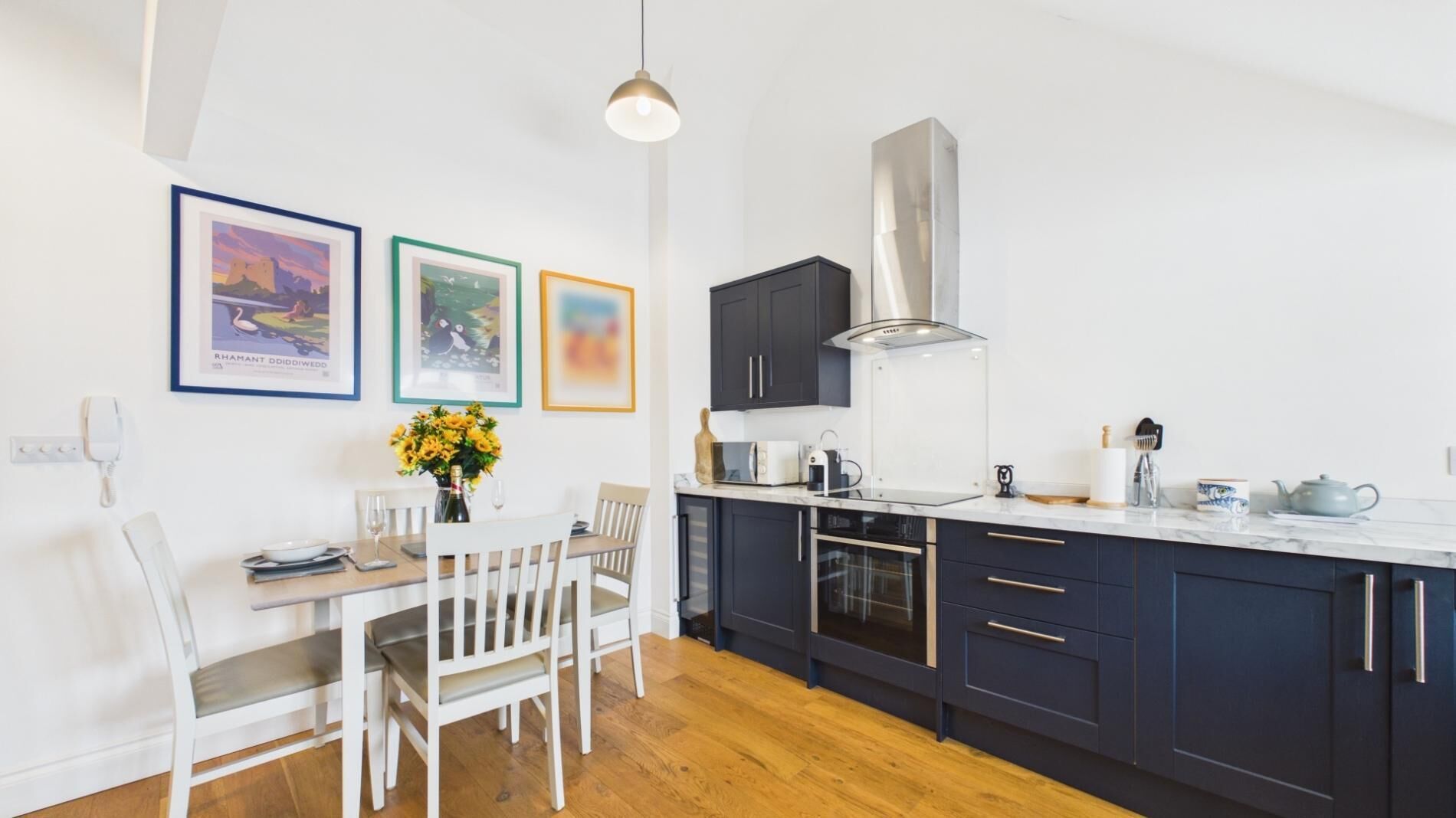 Una cocina moderna con armarios oscuros, una campana extractora de acero inoxidable y una zona de comedor con sillas blancas y una mesa.